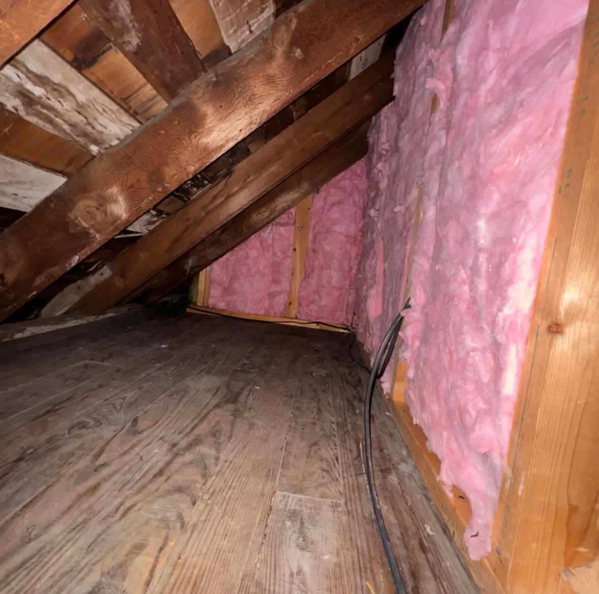 Damaged pink fiberglass insulation in attic with exposed wooden beams and joists